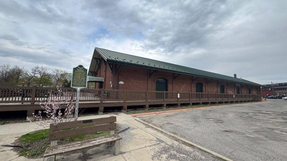 Photo of a brick building with a green roof set behind sidewalk and a parking lot.