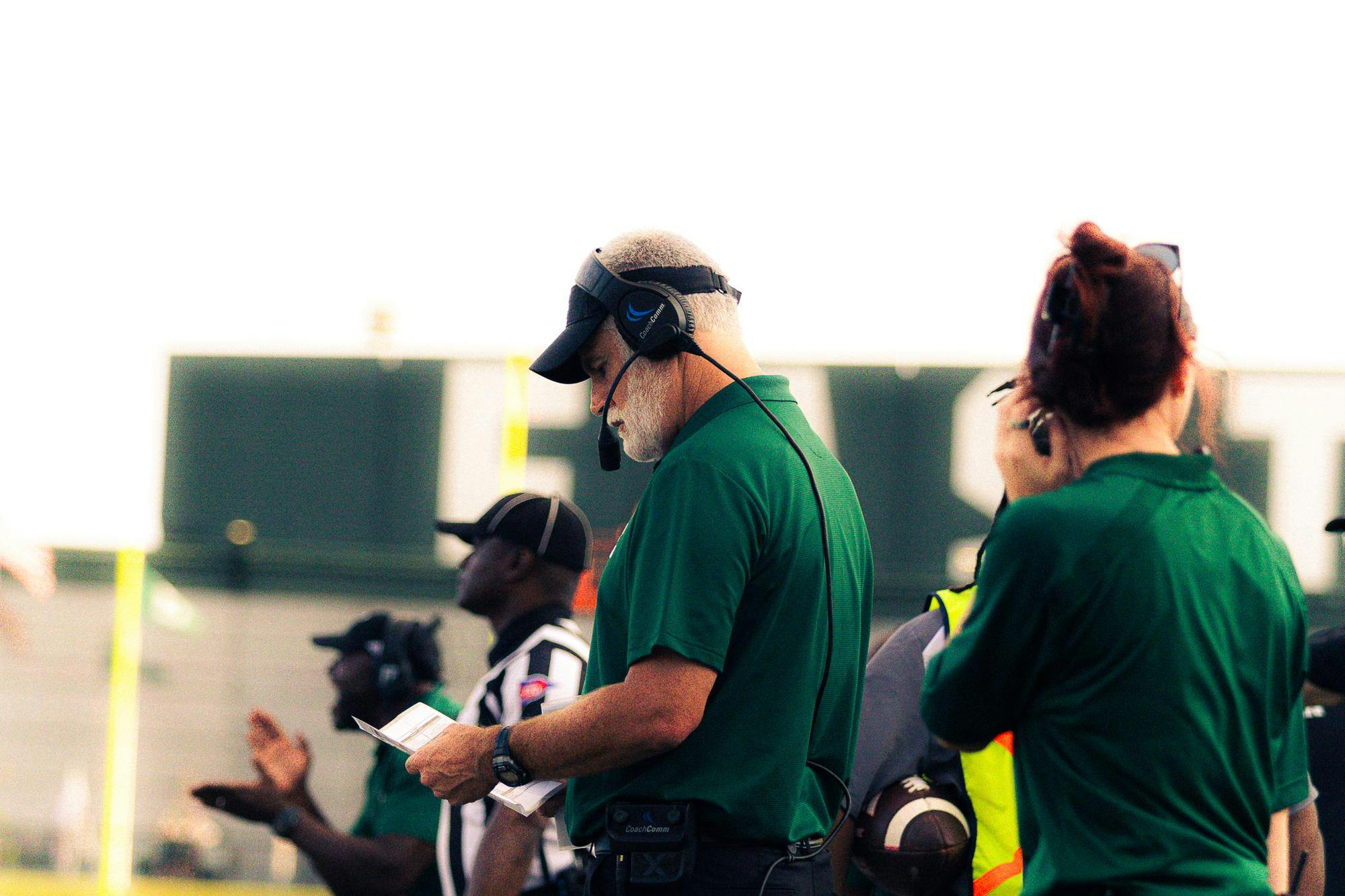 The Eastern Michigan head coach wearing a green polo shirt and a headset looks down at a play sheet during a critical moment in the action.