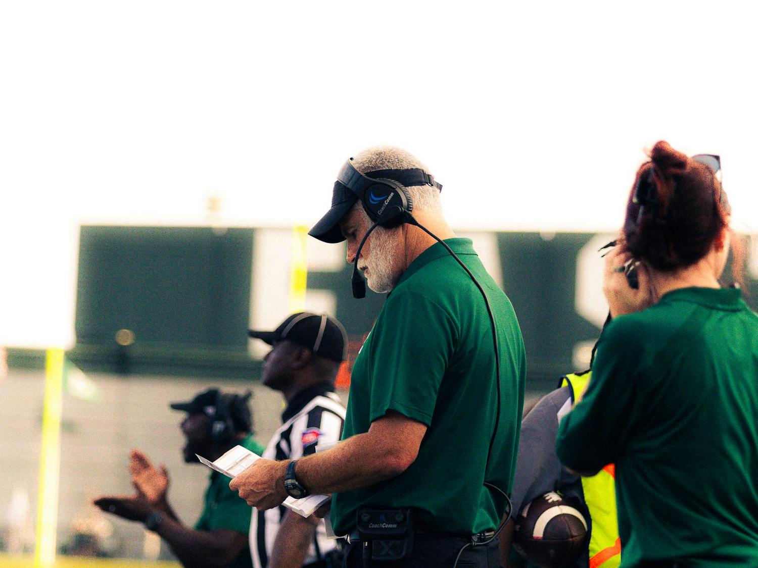 The Eastern Michigan head coach wearing a green polo shirt and a headset looks down at a play sheet during a critical moment in the action.