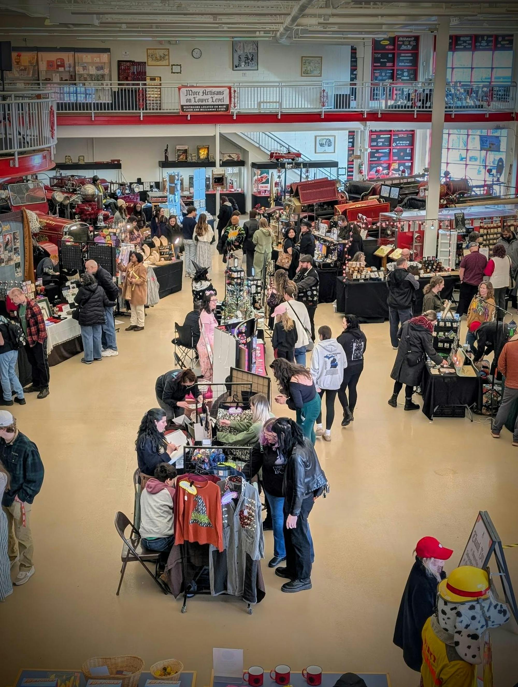 Customers gather around vendors' booths in a large, well-lit room with a beige-colored floor and white walls. The vendors are selling various merchandise, including shirts, jewelry and more.