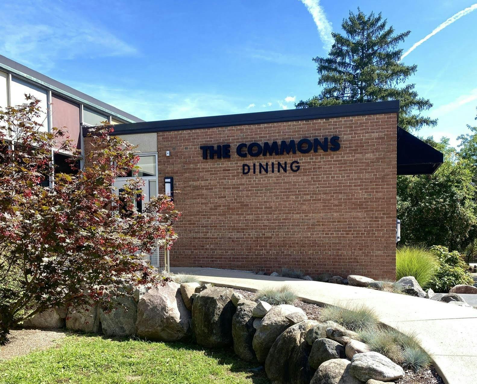 Sidewalk leading up to a metal door on a brick wall with "The Commons Dining" written in black letters.