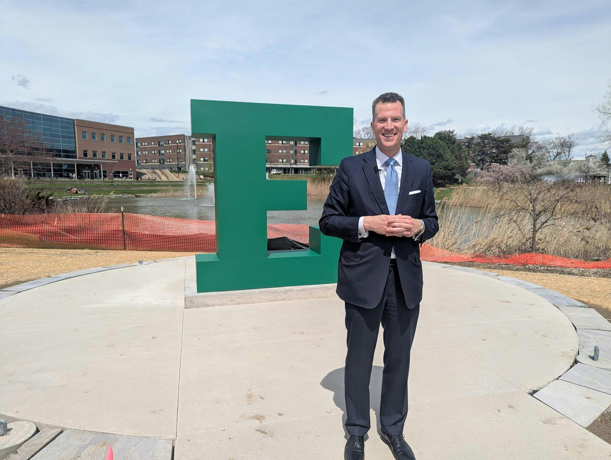Brendan Kelly smiles and stands in front of a large block "E" sculpture, with construction fencing in the background and the university pond in the distance.