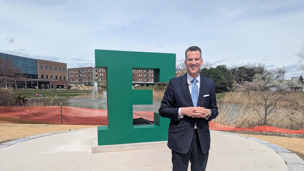 Brendan Kelly smiles and stands in front of a large block "E" sculpture, with construction fencing in the background and the university pond in the distance.