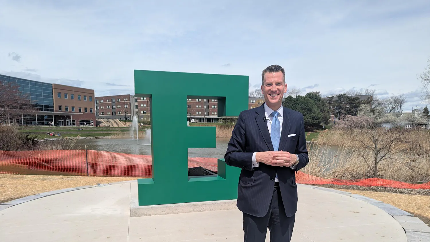 Brendan Kelly smiles and stands in front of a large block "E" sculpture, with construction fencing in the background and the university pond in the distance.
