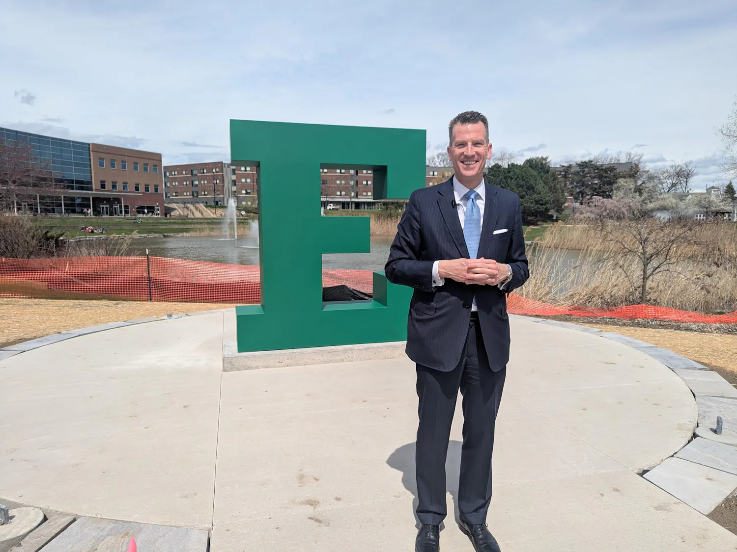 Brendan Kelly smiles and stands in front of a large block "E" sculpture, with construction fencing in the background and the university pond in the distance.