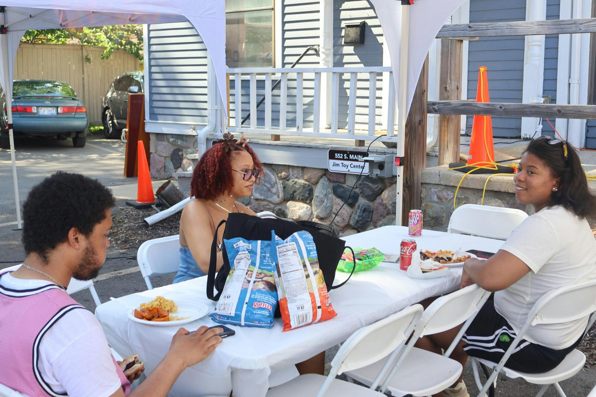 Three people are sat down at a folding table each with a plate of food. There are two bags on chips on the table.