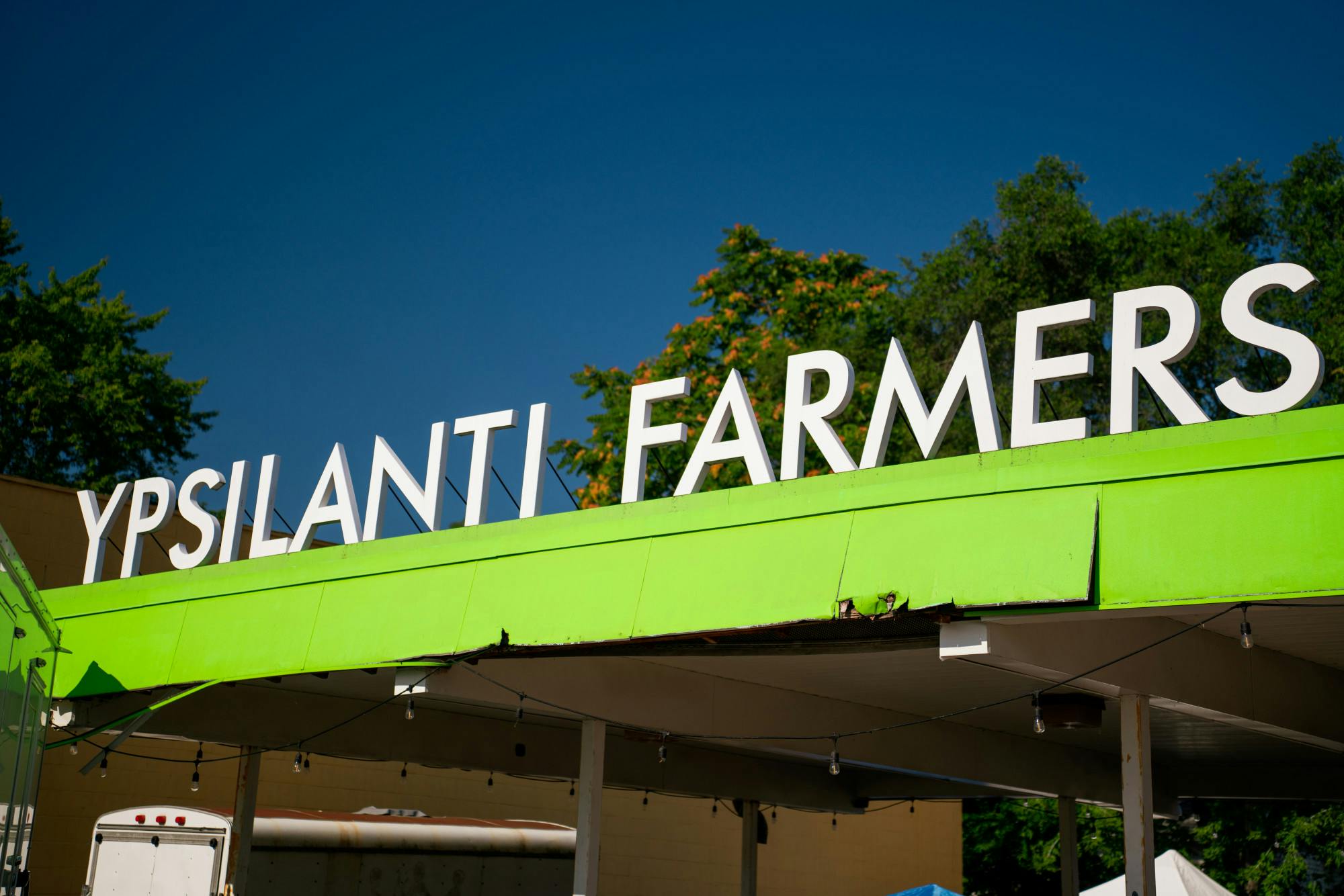 White letters, spelling out Ypsilanti Farmers, line the top of a bright yellow canopy over the area that is home to the Ypsilanti Farmers Market. 