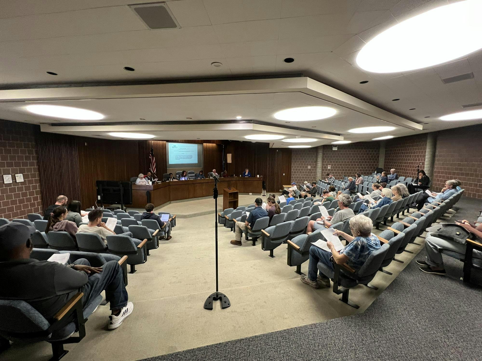 A wide-angle shot of a medium-sized room with rows of chairs that are sporadically filled with attendees. In the front of the room, eight trustees sit behind a long wooden desk. Three microphones for public comments are visible around the room.