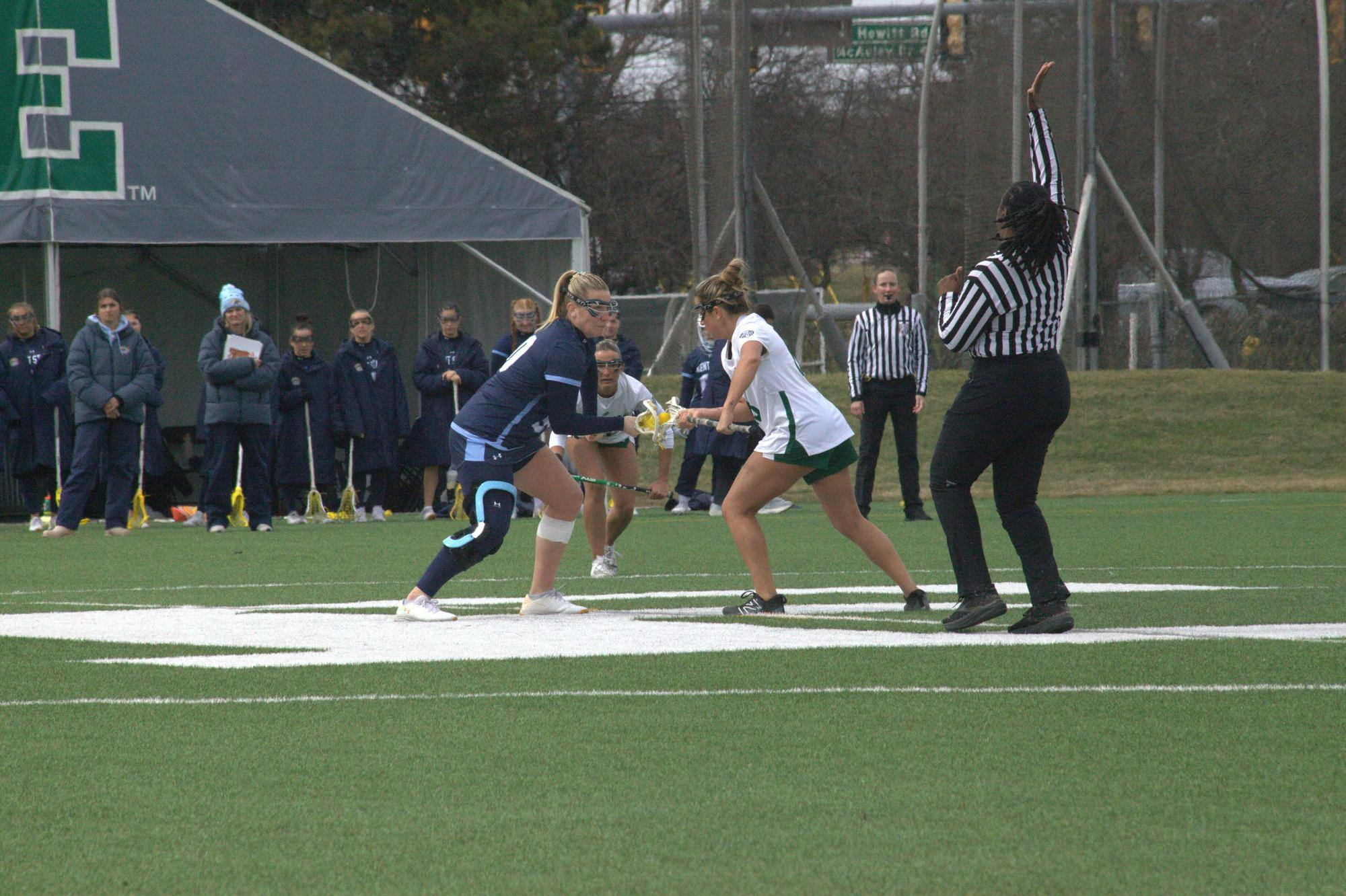 Two women's lacrosse player's of opposing teams face off midfield before a play on an outdoor lacrosse field with a referee off to the side blowing a whistle.