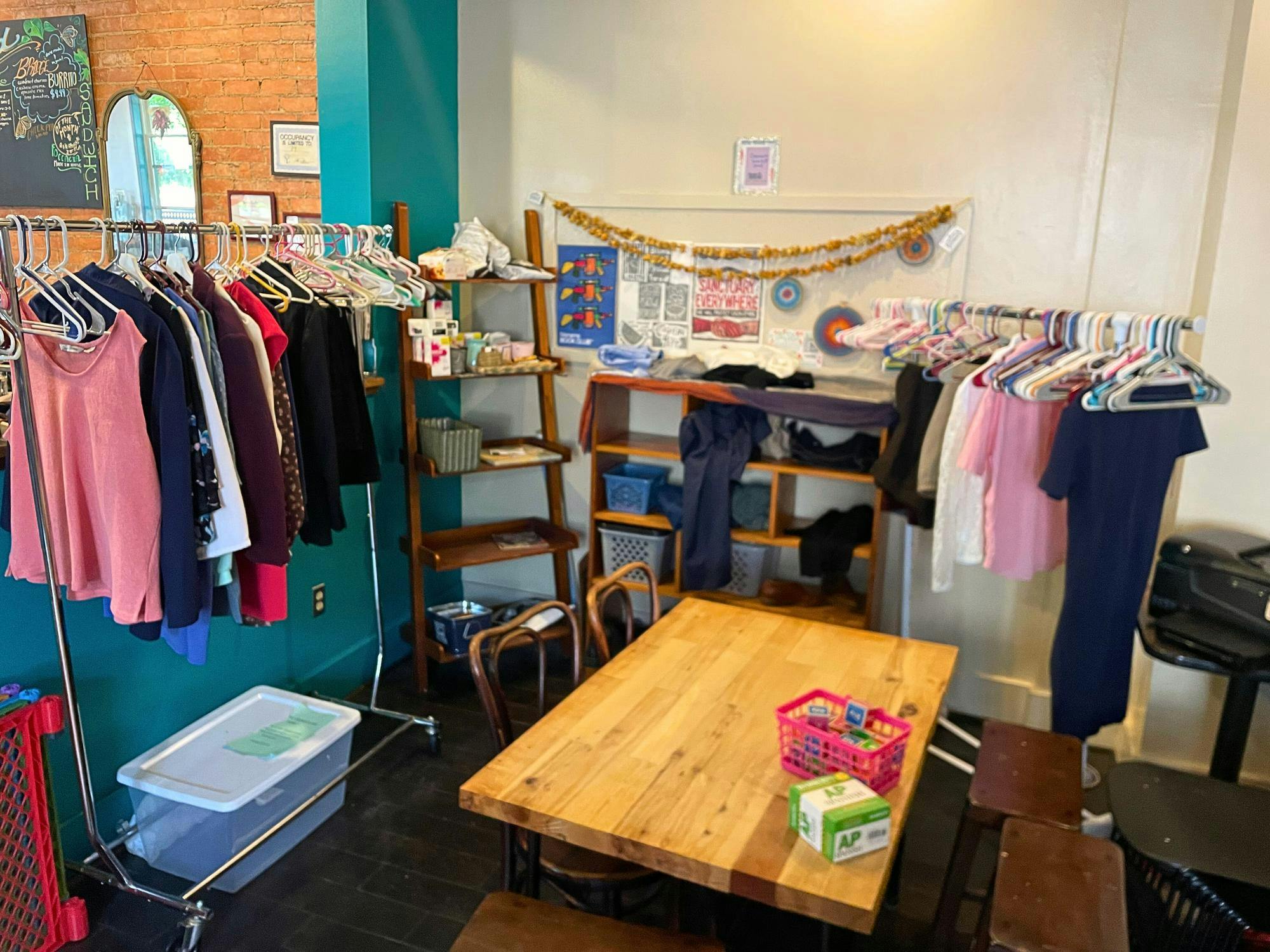 Two clothing racks, two shelves, and a decorated board line the walls in a corner of Bridge Community Cafe. In front of them is a wooden table with chairs and stools that has a basket of activity items sitting on top.