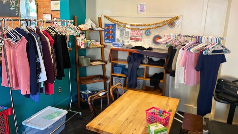 Two clothing racks, two shelves, and a decorated board line the walls in a corner of Bridge Community Cafe. In front of them is a wooden table with chairs and stools that has a basket of activity items sitting on top.