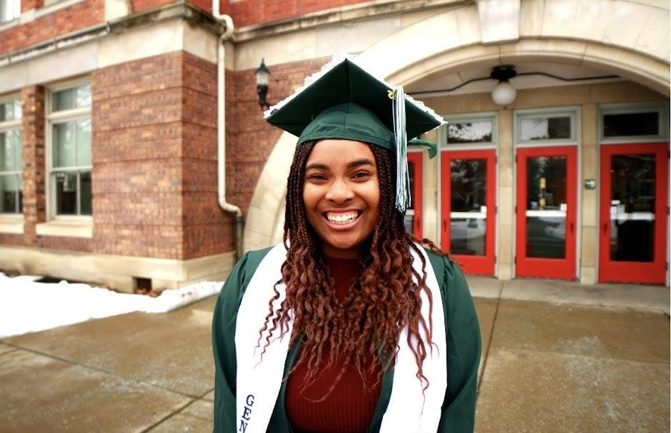 Paris Stinson poses outside in a green graduation cap and gown. A red brick building with a red door stands in the background.