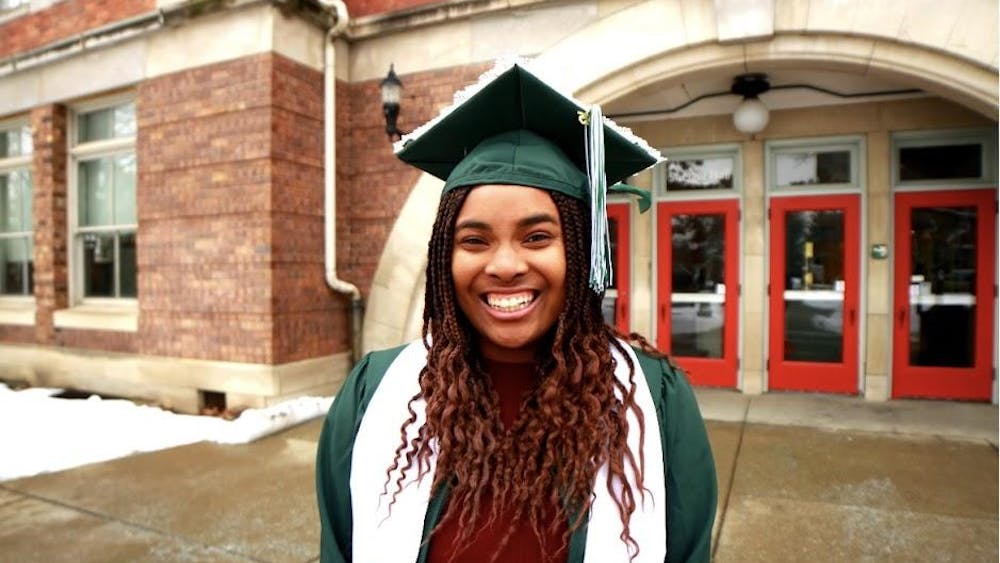 Paris Stinson poses outside in a green graduation cap and gown. A red brick building with a red door stands in the background.