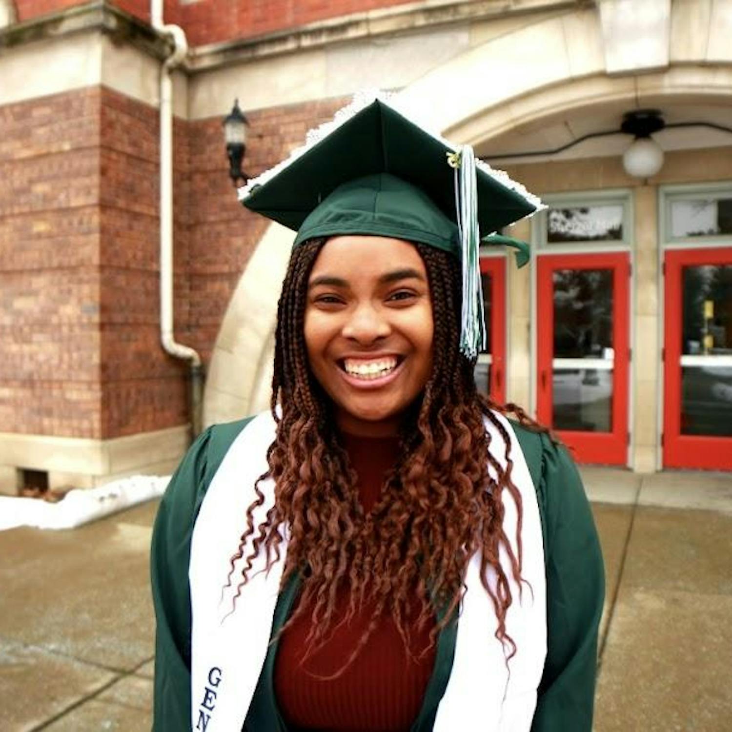 Paris Stinson poses outside in a green graduation cap and gown. A red brick building with a red door stands in the background.