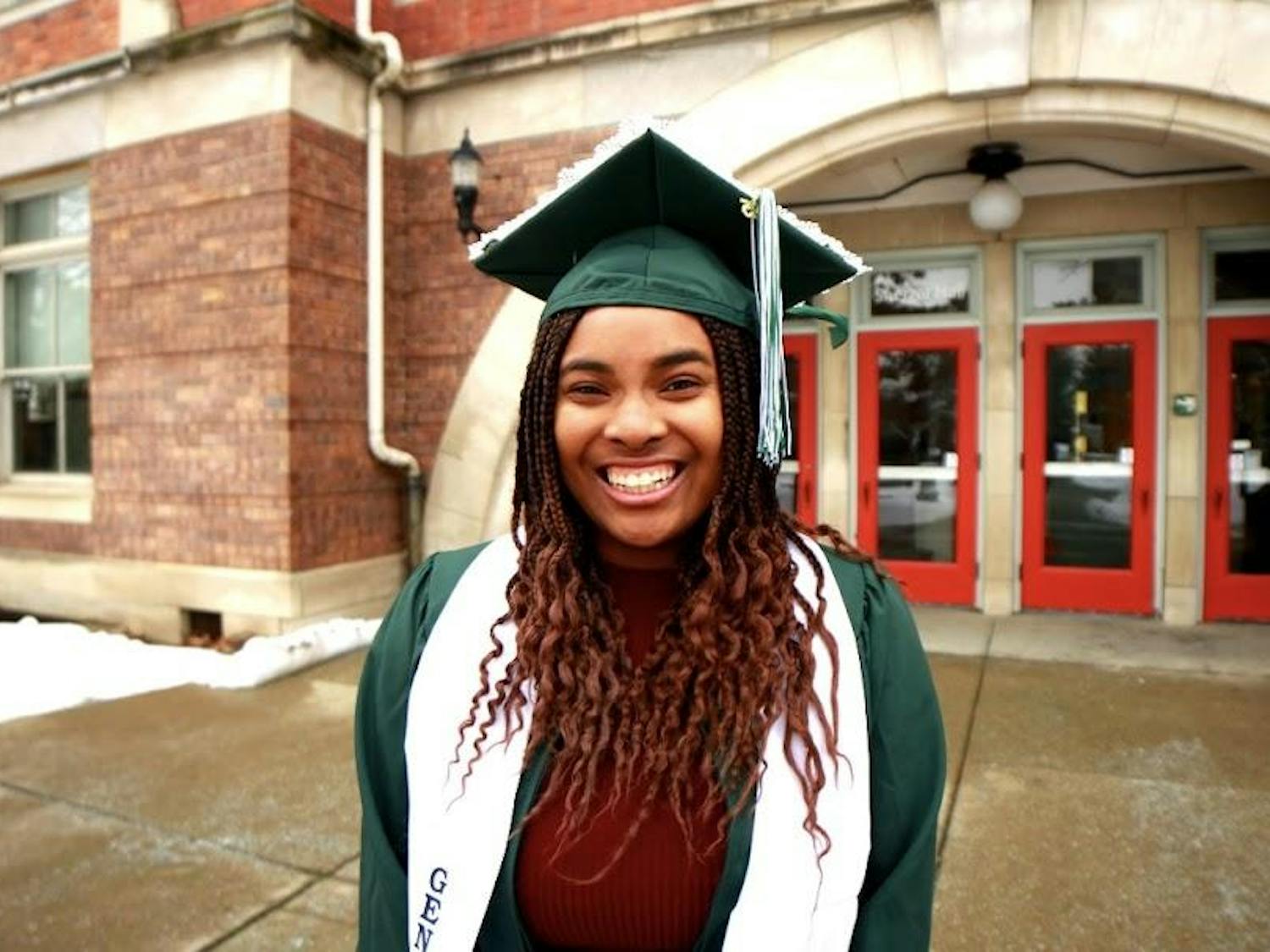Paris Stinson poses outside in a green graduation cap and gown. A red brick building with a red door stands in the background.