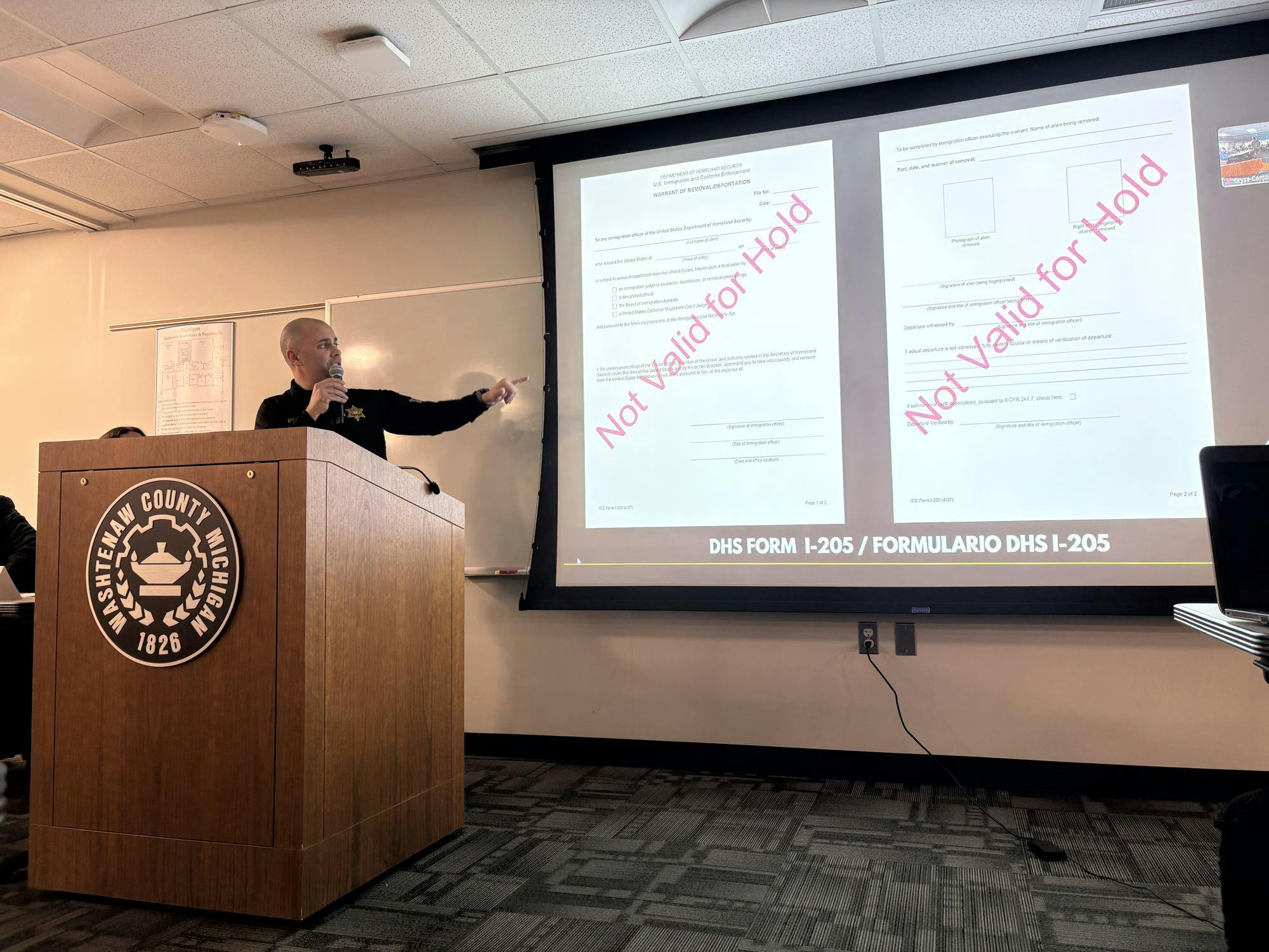 Kurt Schiappacasse stands in front of a projector displaying two types of immigration documents, a detainer and a warrant for removal/deportation. The documents have large red text over them that say "Not Valid for Hold."