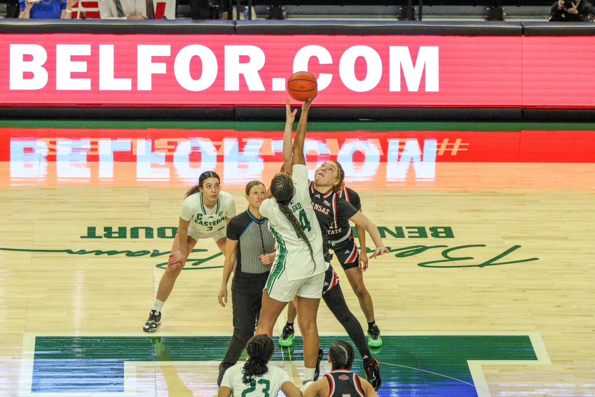 Two players from each team reach for the basketball after it is tossed high up in the air. A referee and an additional player from each team watch.