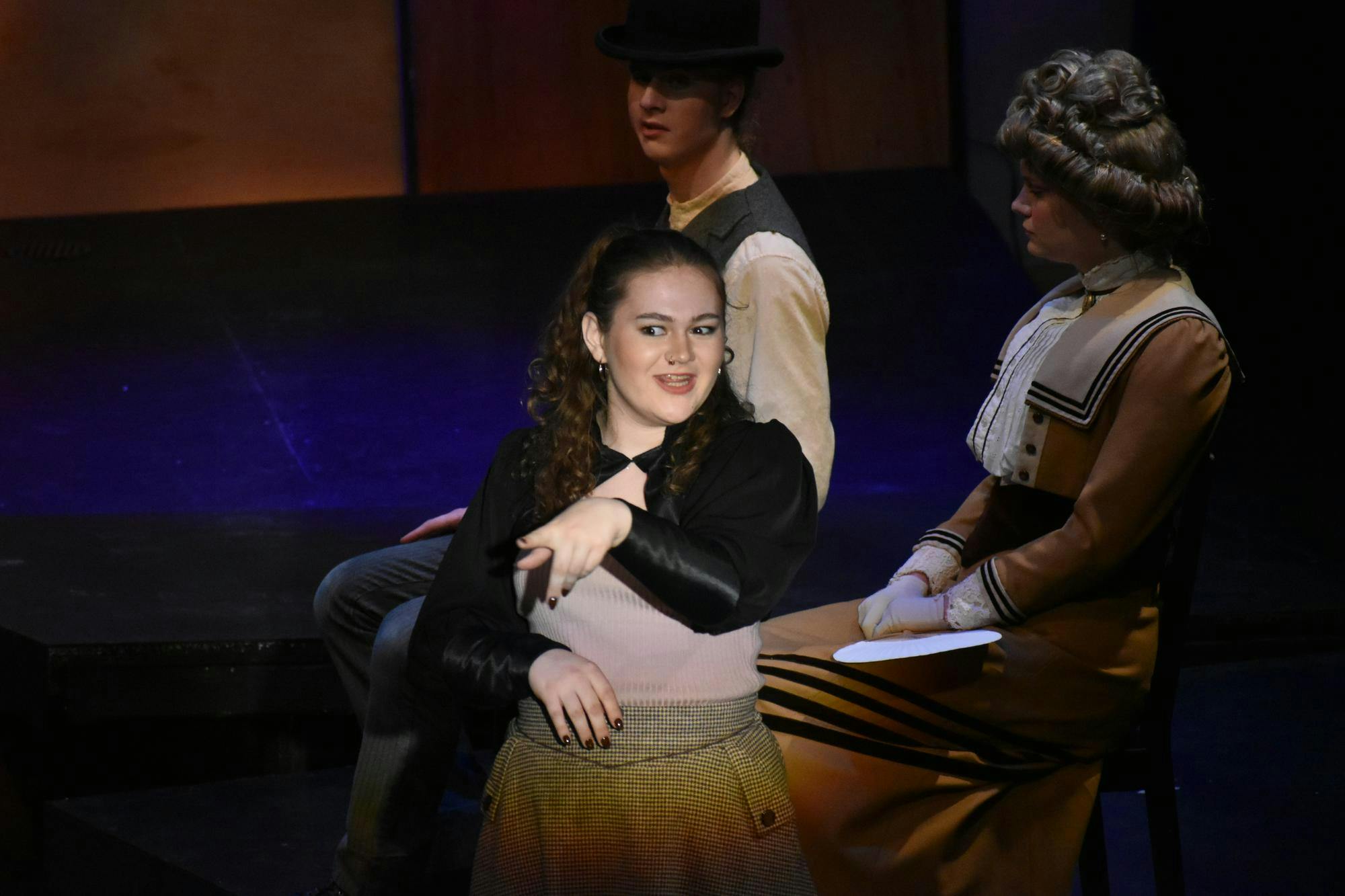 An actor stands center stage in the spotlight, pointing to the audience. Two actors sit in the background.