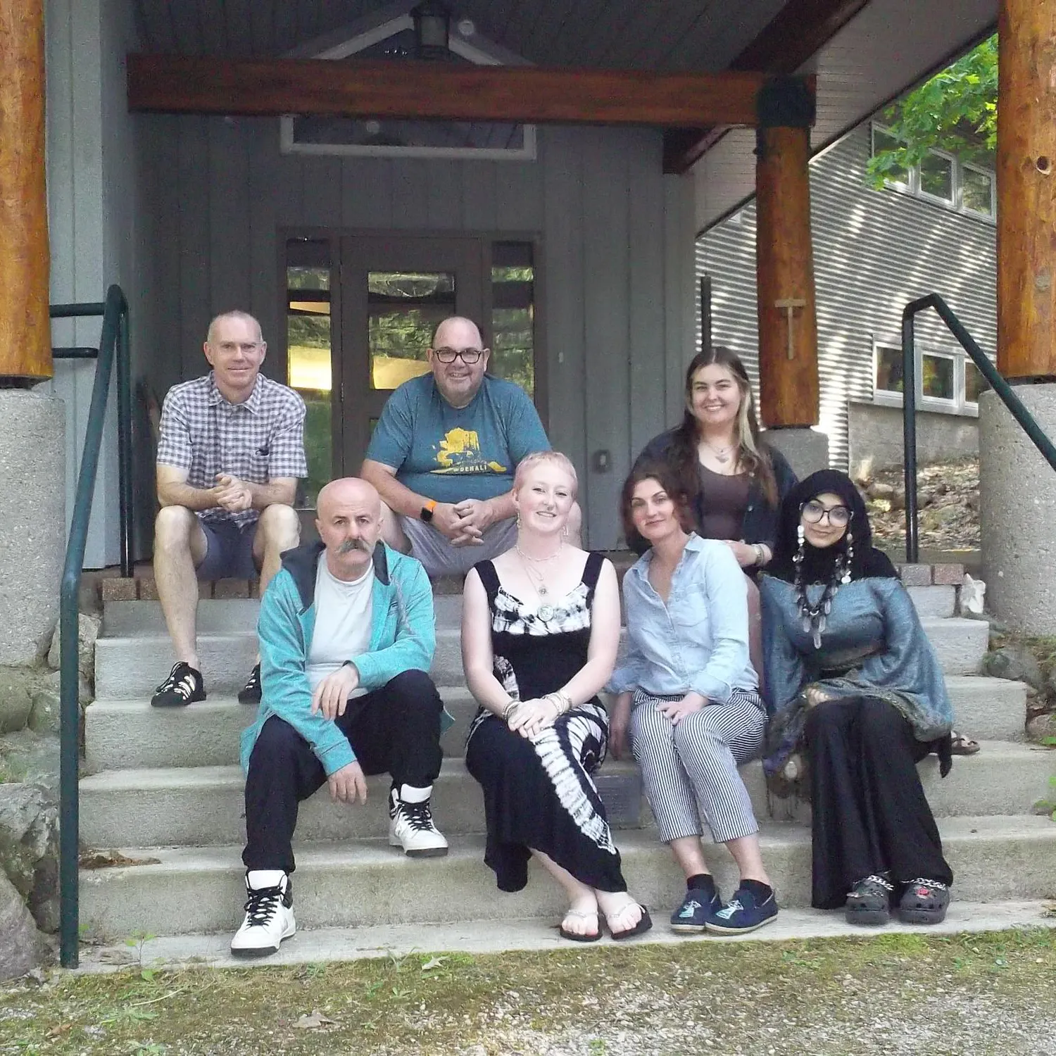 7 adults sit on concrete stairs outdoors in front of a gray building.