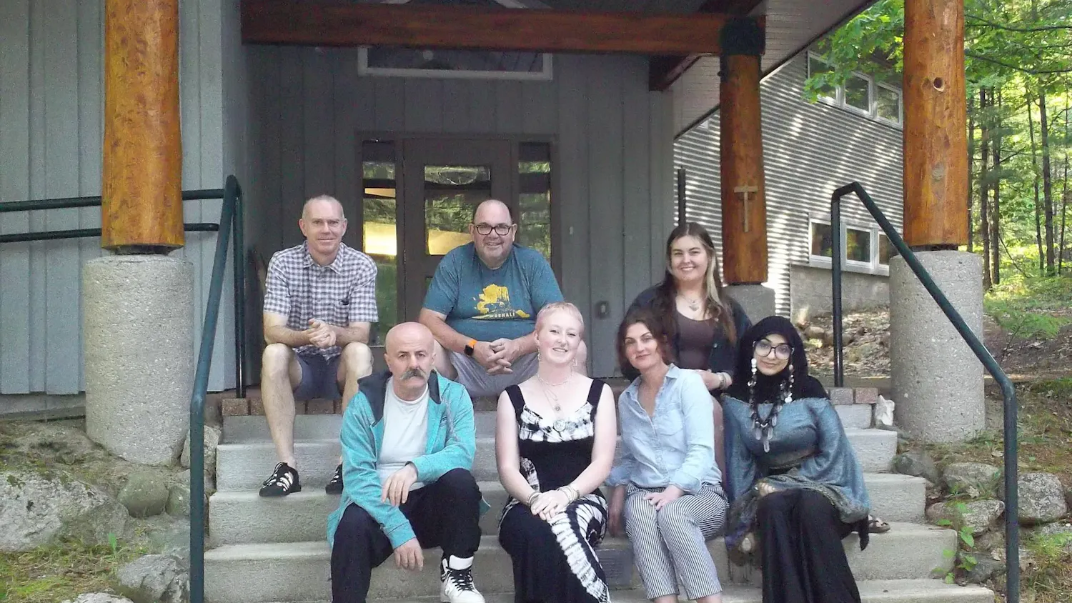7 adults sit on concrete stairs outdoors in front of a gray building.