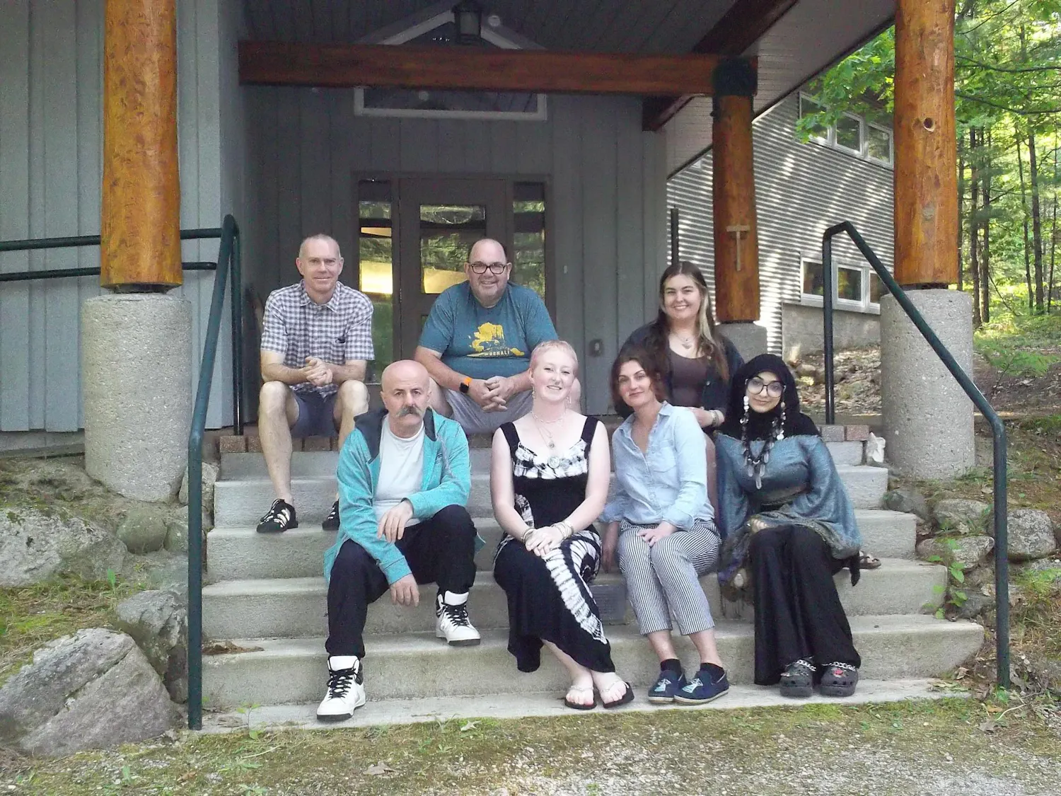 7 adults sit on concrete stairs outdoors in front of a gray building.