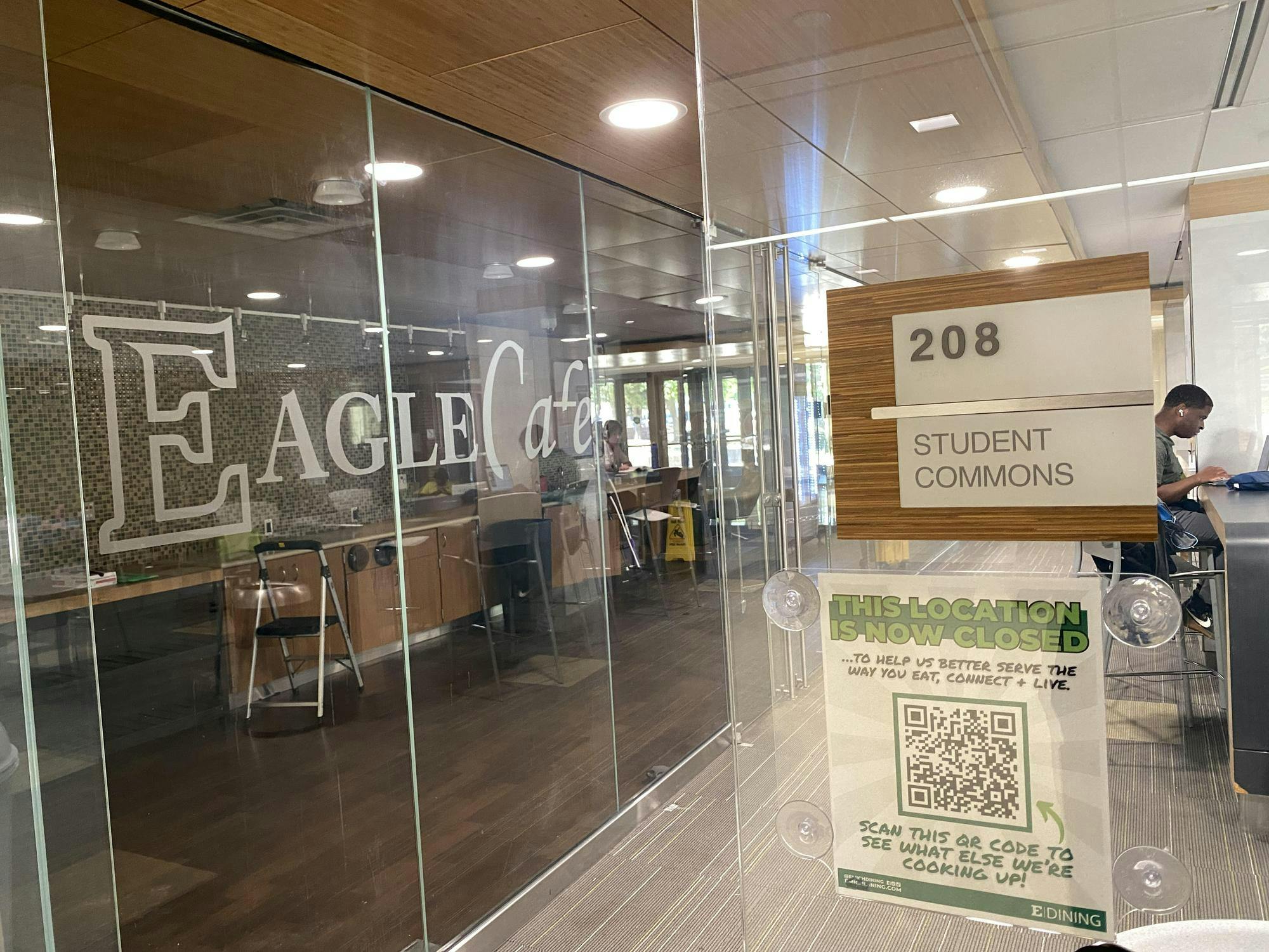 An empty room lined with cabinets and countertops against the back wall is visible behind a glass wall with the words, "Eagle Cafe." A sign in the foreground reads, "This location is now closed."
