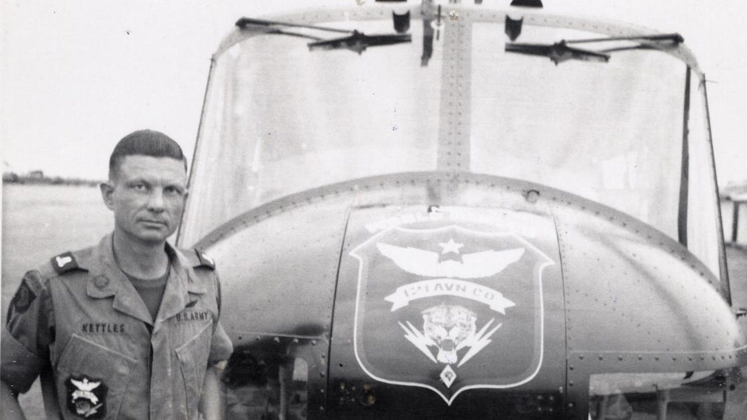 In a black and white photo, Charles Kettles, dressed in an army uniform, stands in front of a helicopter.