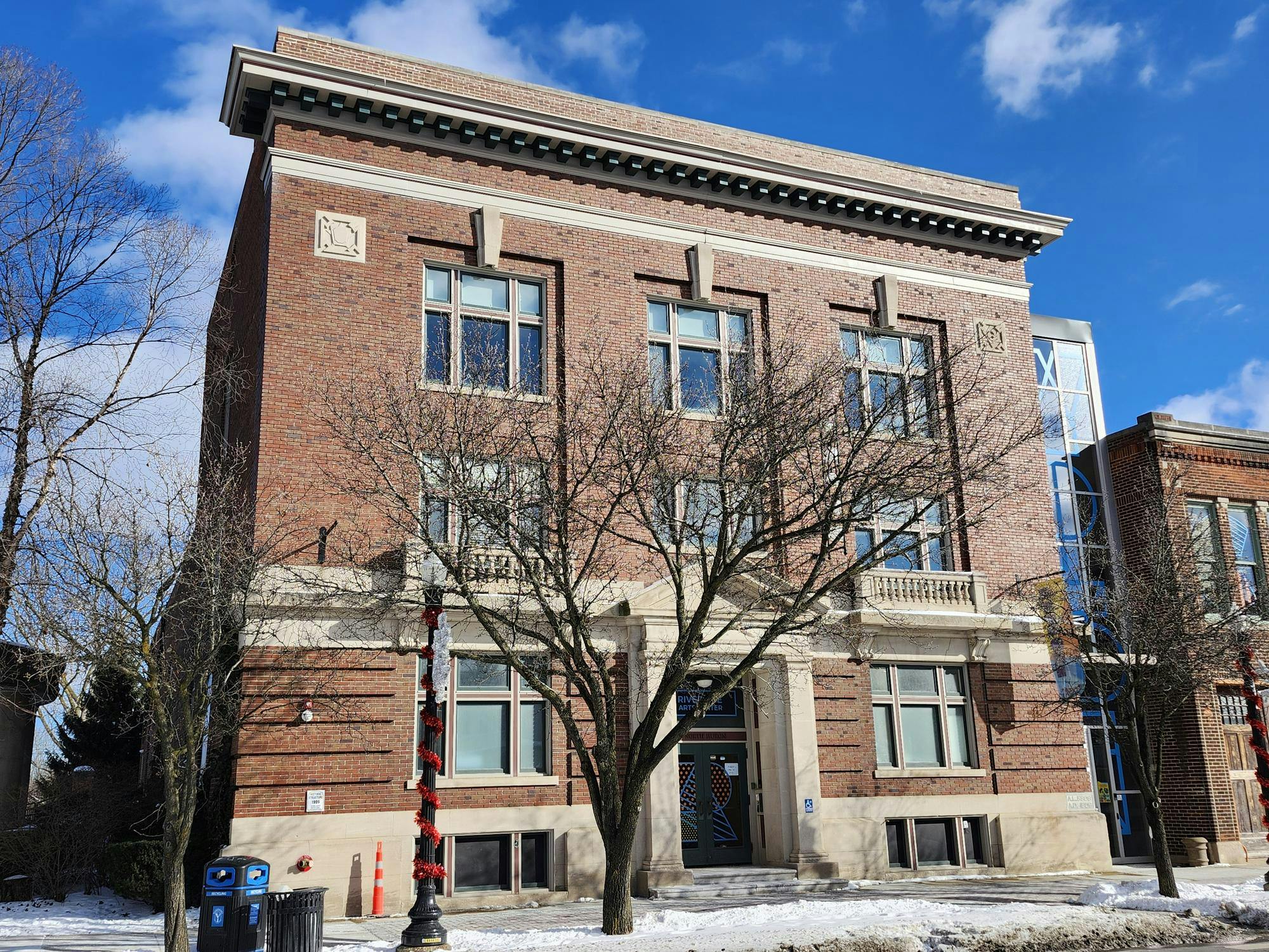 A brick building with a lampost and spaced-out bare trees in the foreground in the snow. The sky is lightly cloudy in the background. Another brick building stands to the right of the first.