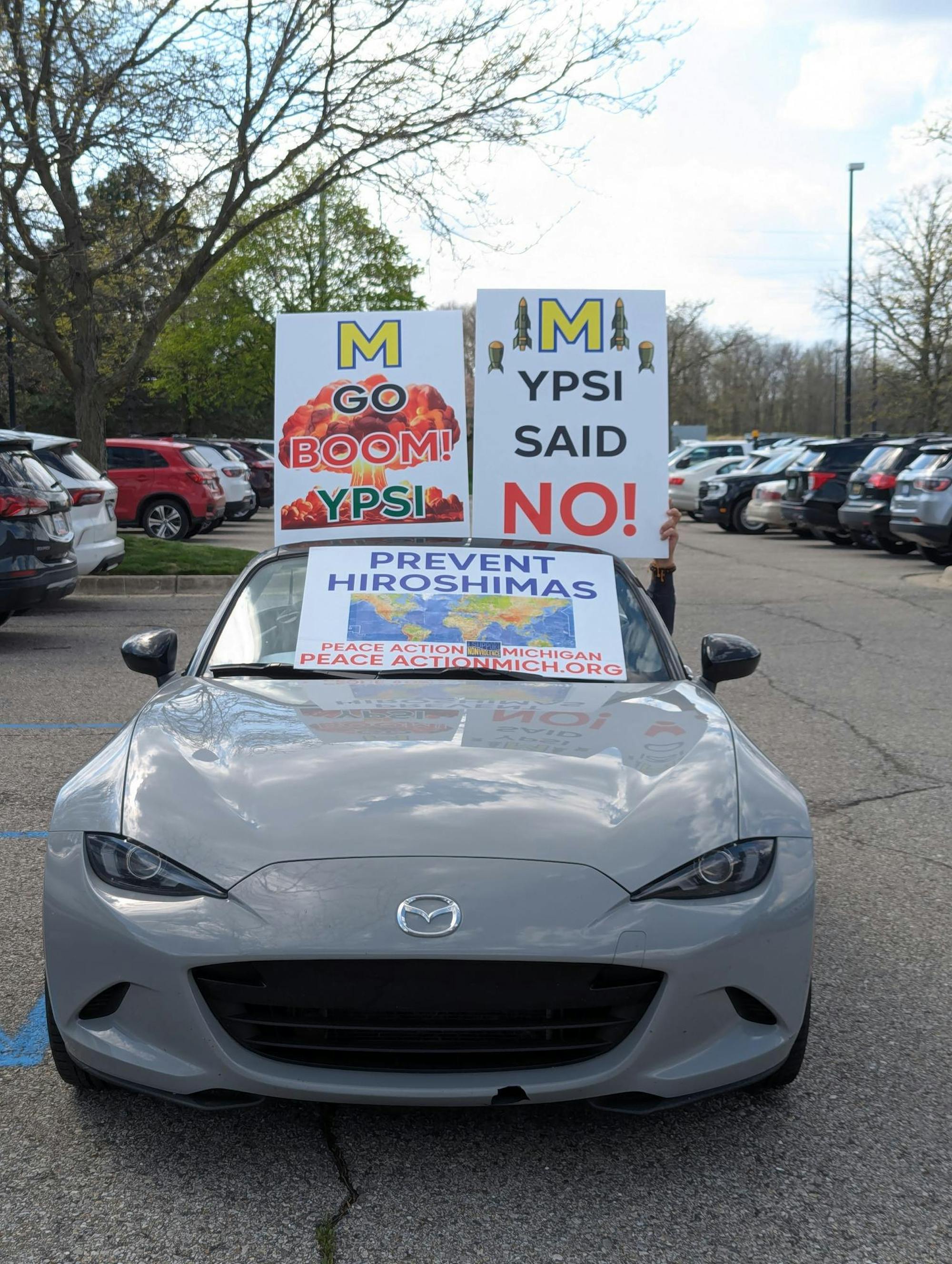 A car is in a parking lot and a driver, whose face is not shown, holds up and displays signs that say "M Go Boom! Ypsi" and "M Ypsi said no!" with the University of Michigan Block M and "Prevent Hiroshimas, Peace Action Michigan."