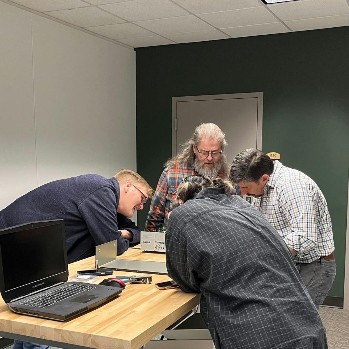 4 people lean over a wooden table to fix electronic equipment.