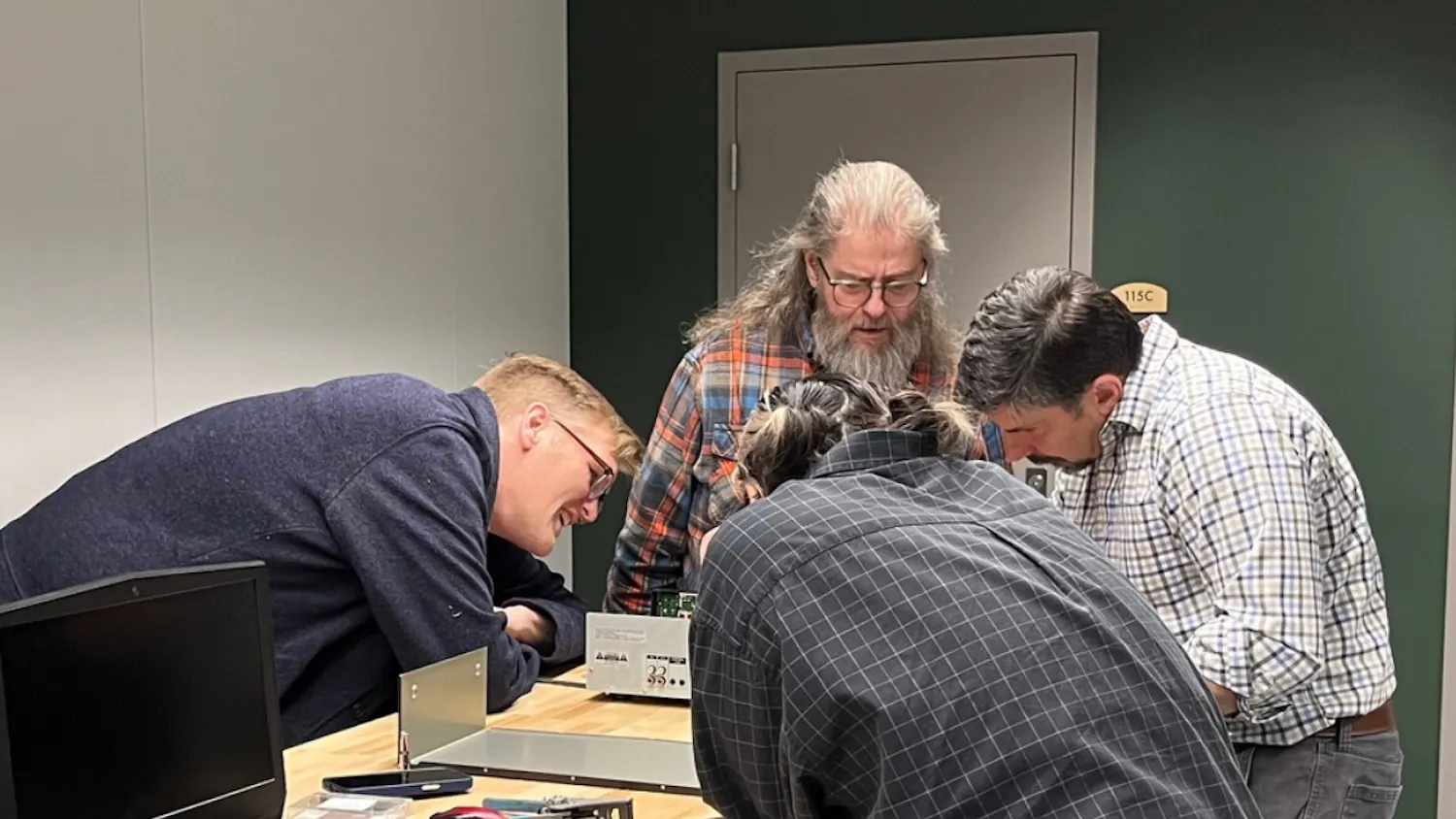 4 people lean over a wooden table to fix electronic equipment.
