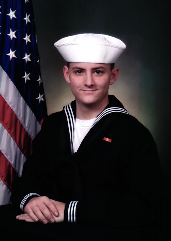 John Andrew Jenuwine, from the waist up, in a military Navy uniform, sitting next to an American Flag.