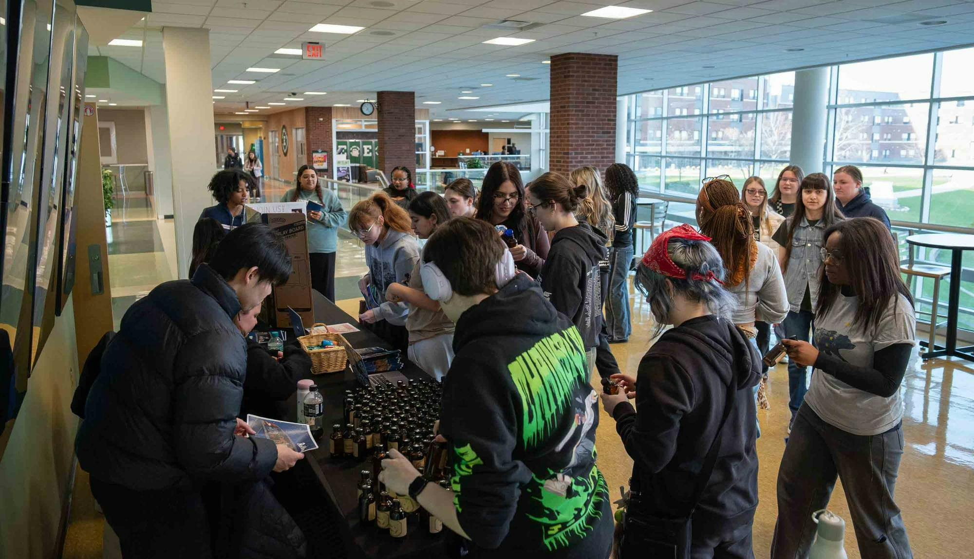 About 20 students surround a black table full of small glass bottles in a brightly lit, modern styled room.
