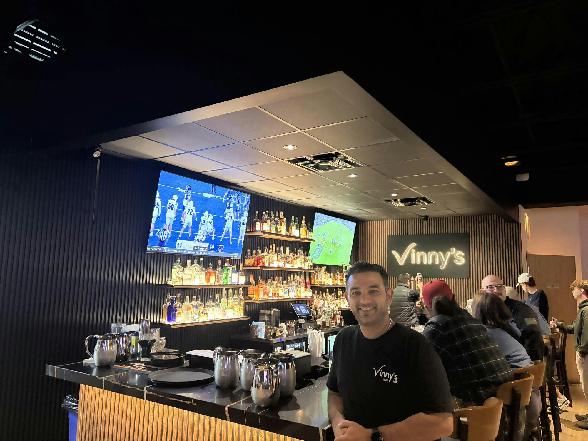 Vince Ankawi stands in front of a bar lined with bottles of liquor; patrons sit on bar seats behind him while a football game plays on a TV screen in the background.