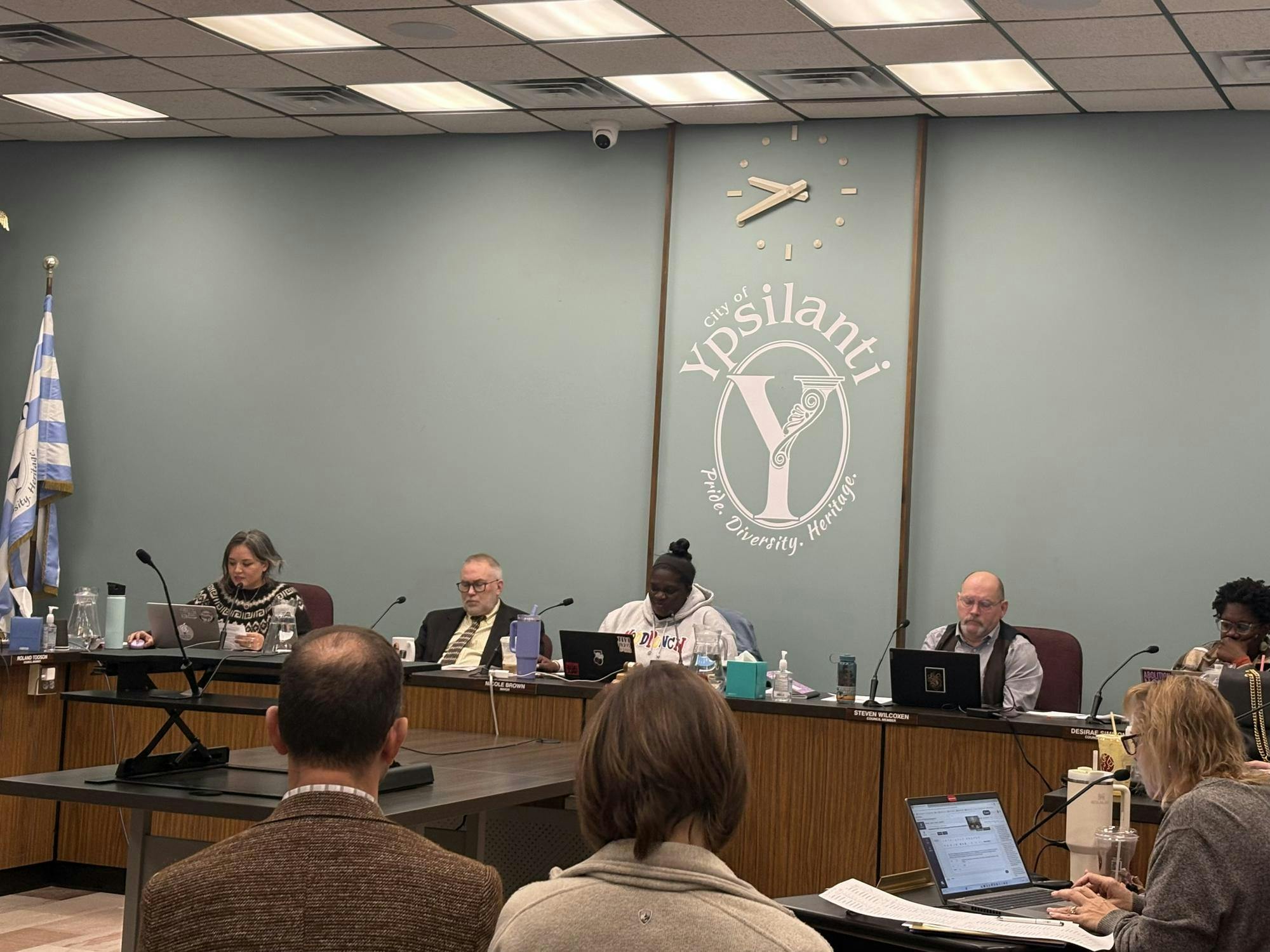 Six Ypsilanti City Council members sit at wooden desks with laptops and microphones in front of them. The logo for the City of Ypsilanti is painted on the wall behind them.