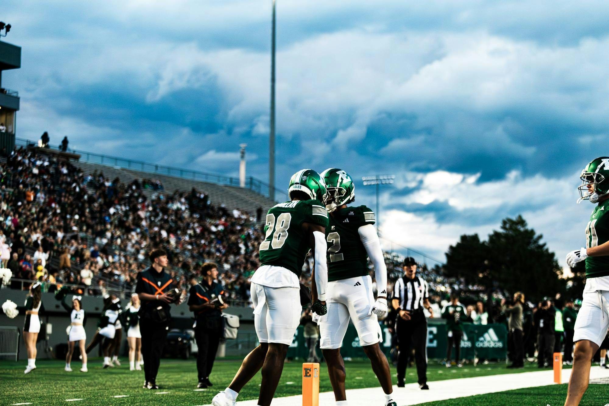 Two EMU football players touch green helmets together in celebration in front of the crowd and a gray, cloudy sky.
