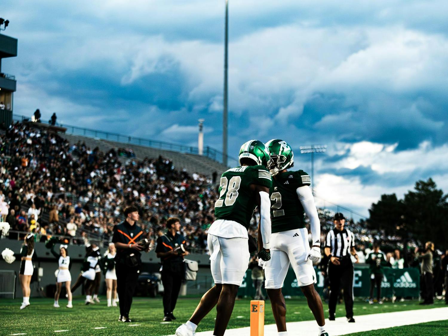 Two EMU football players touch green helmets together in celebration in front of the crowd and a gray, cloudy sky.