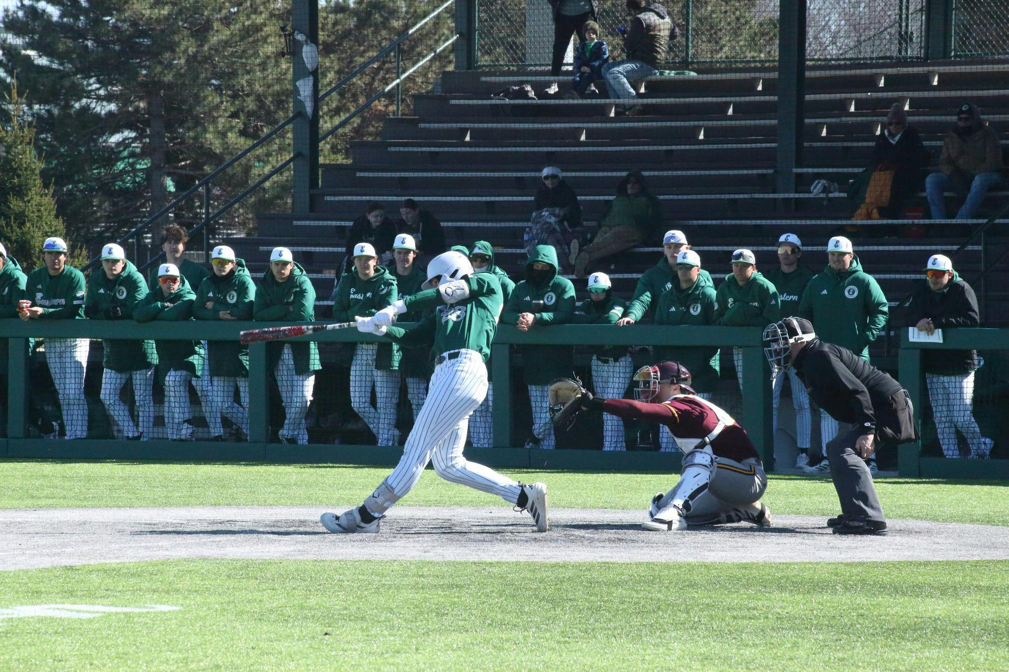 A baseball player mid-swing at outdoor game. Teammates stand in the background.