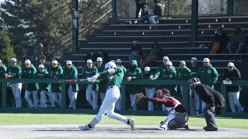 A baseball player mid-swing at outdoor game. Teammates stand in the background.