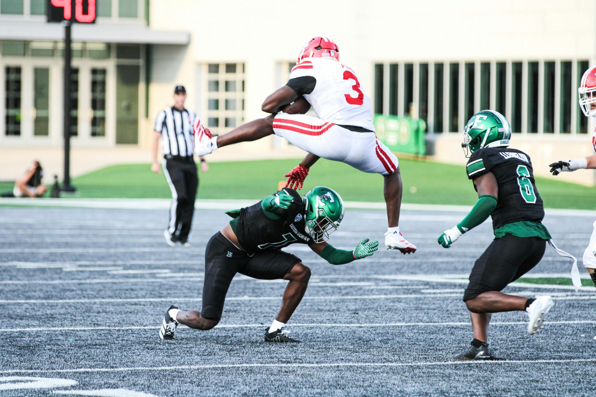A Louisiana Lafayette player decked out in red and white jumps airborne over an Eastern Michigan defender wearing black and green with a football in hand. 