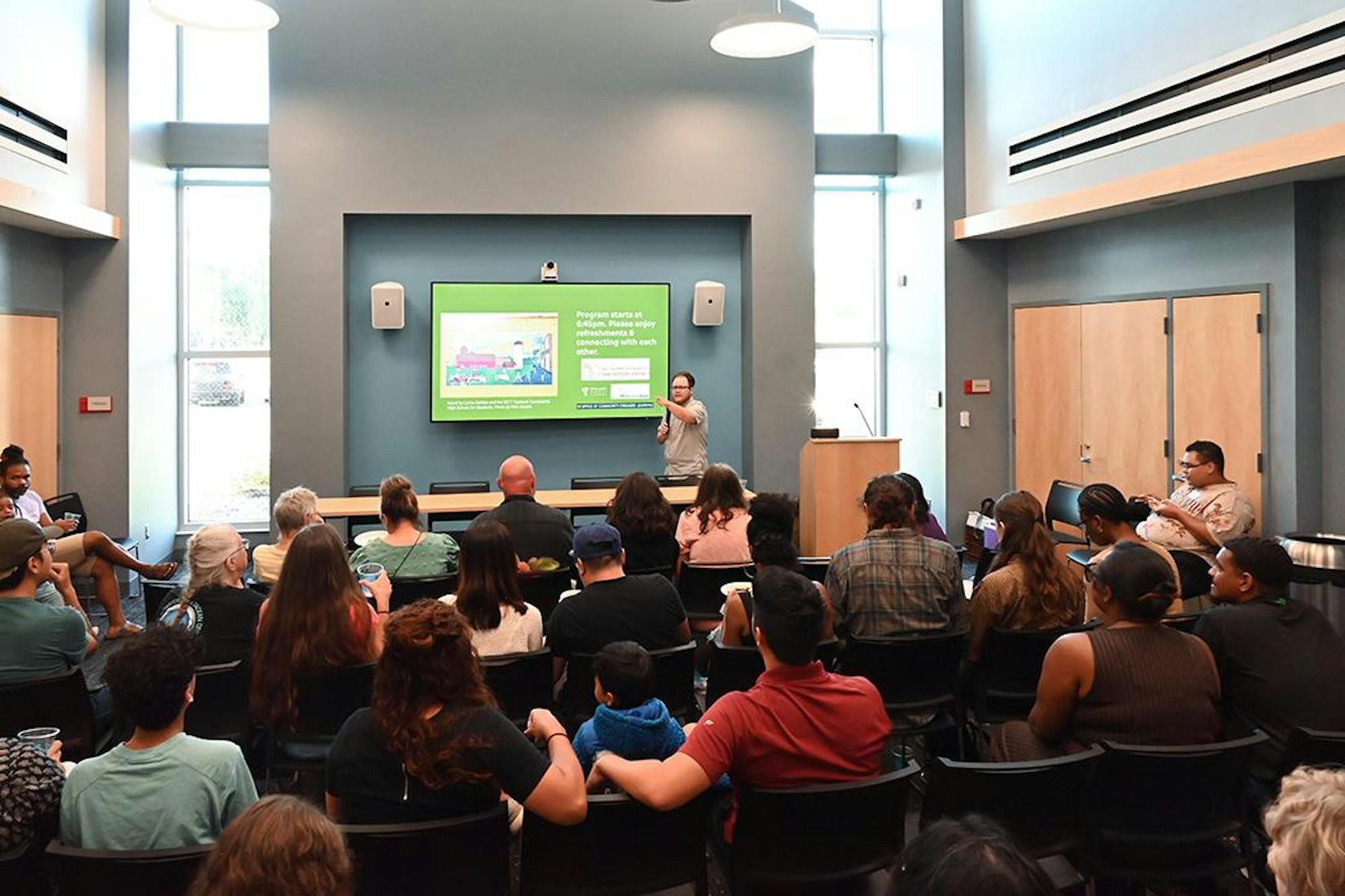 The backs of a crowd are seen as a man addresses the group and displays a presentation.