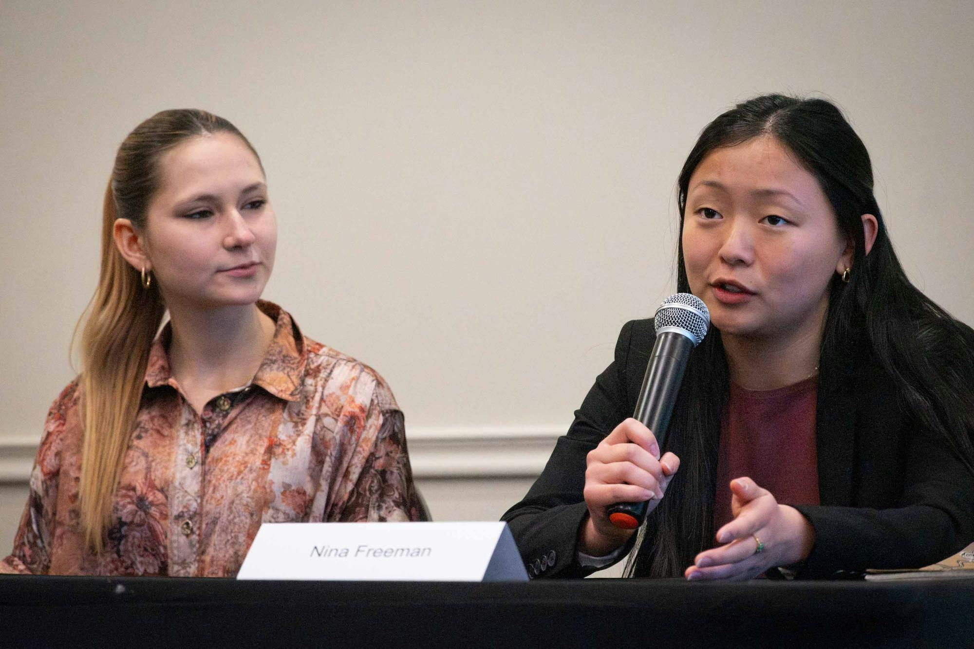 Eva Orlich Rojas and Nina Freeman sit at a table during the candidate forum. Freeman speaks into a microphone.