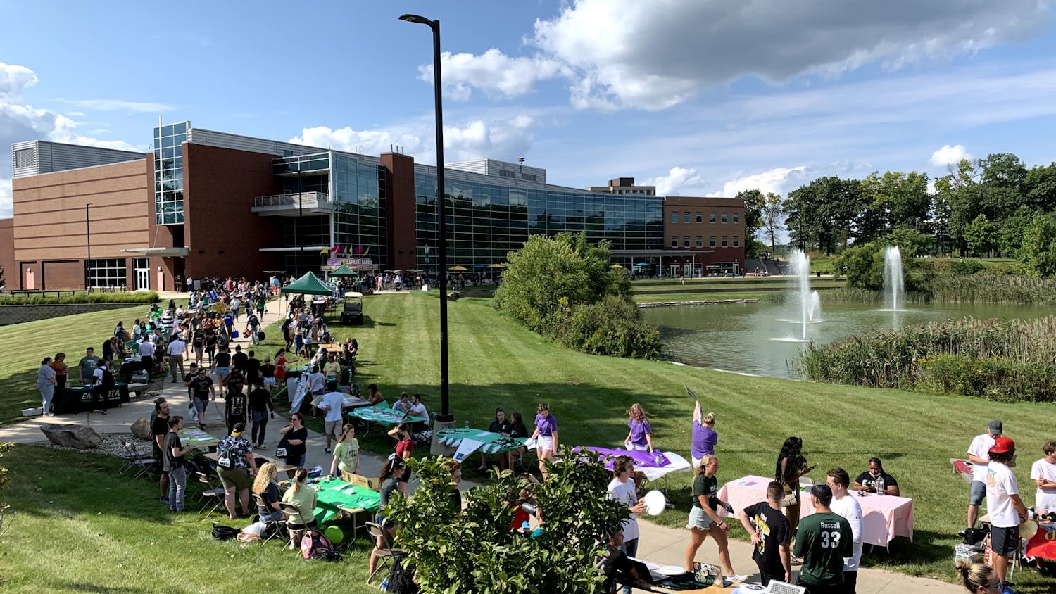 Tables from varying student organizations line the sidewalk in front of the Student Center. Many students walk the sidewalk between the tables.