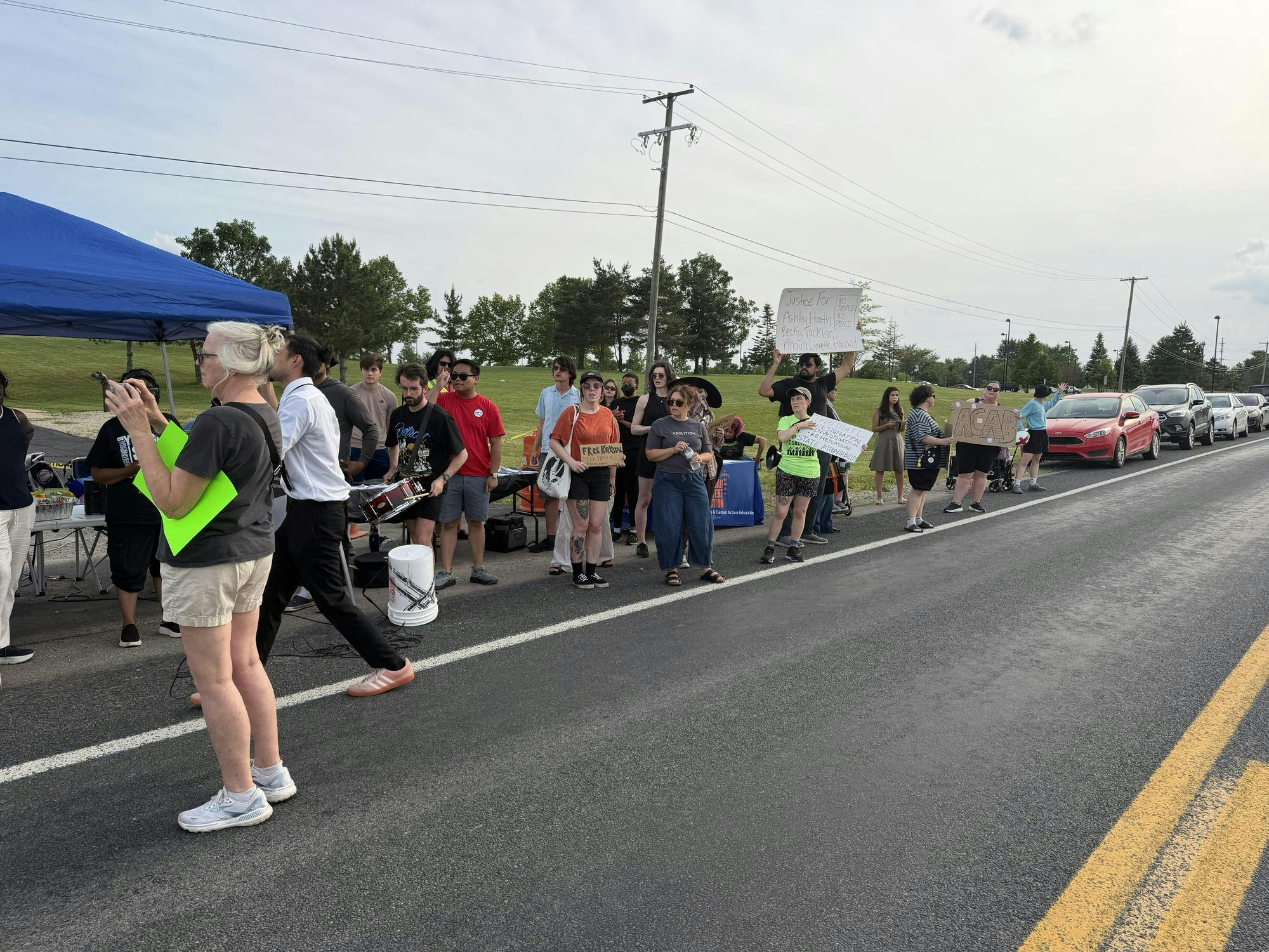 People hold signs on the side of the road in front of power lines and trees. 