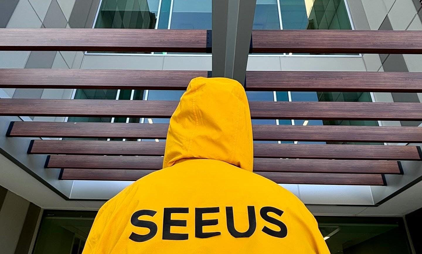 A student in a bright yellow jacket stands under the entrance to an EMU building, the word "SEEUS" displayed on the back of the jacket in black. The worker's hoodie is up, and their back faces the camera.