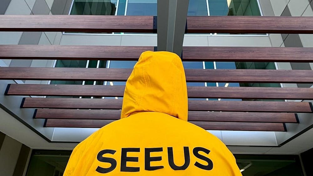 A student in a bright yellow jacket stands under the entrance to an EMU building, the word "SEEUS" displayed on the back of the jacket in black. The worker's hoodie is up, and their back faces the camera.