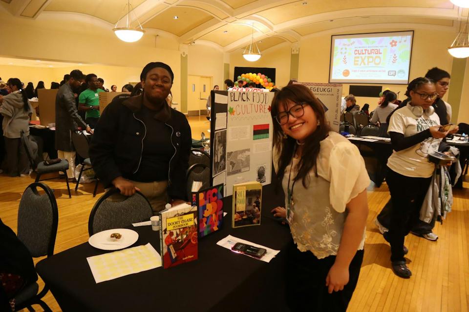 One student stands behind the table, with a poster presentation displayed that says, "Black food culture," and another student stands on the other side of the table. Both are smiling for the photo.
