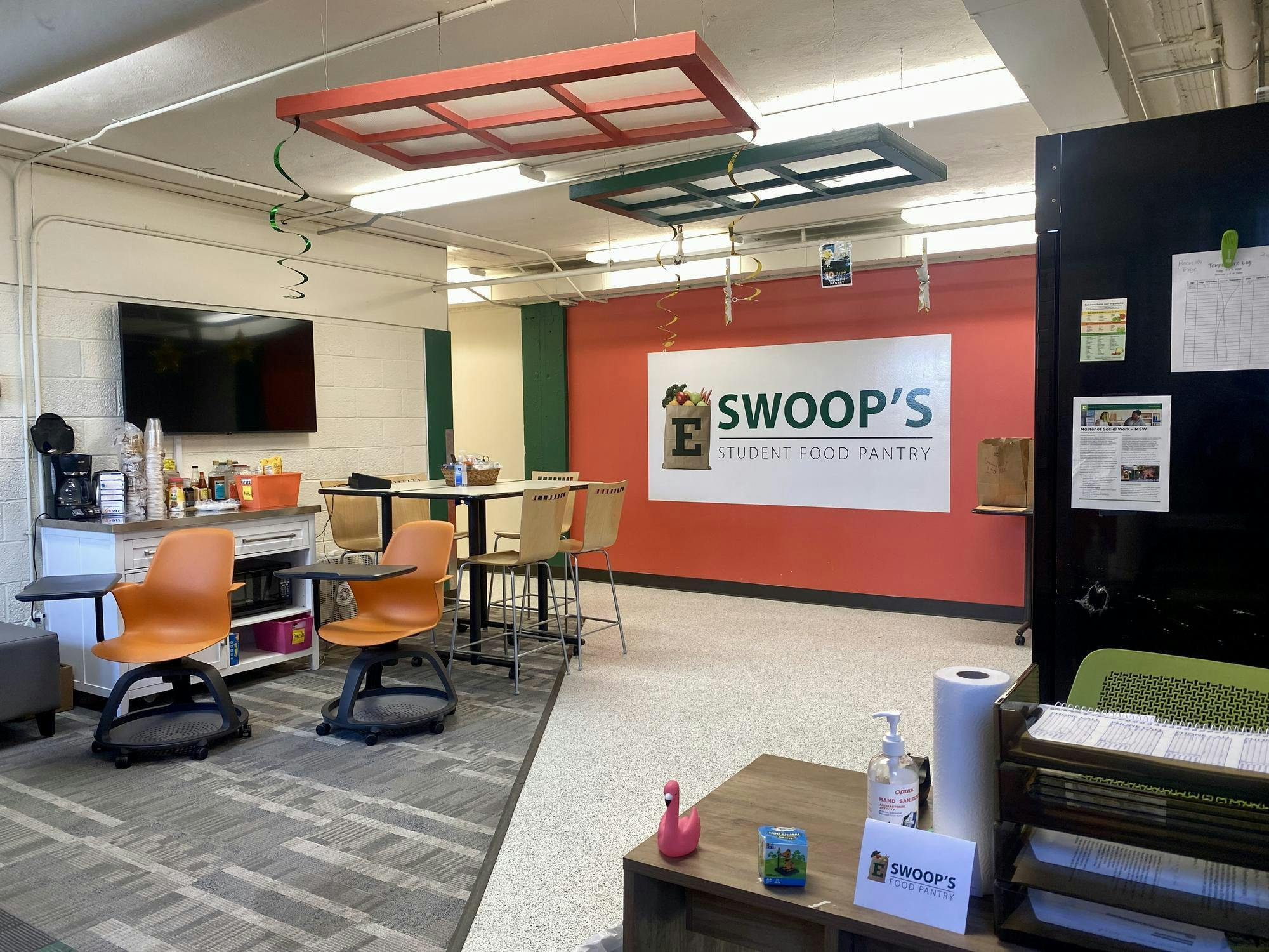 Room with a tall table and four chairs, two rolling chairs, a wall-mounted television and a coffee bar. The back wall of the room is painted orange and reads "Swoop's Student Food Pantry."