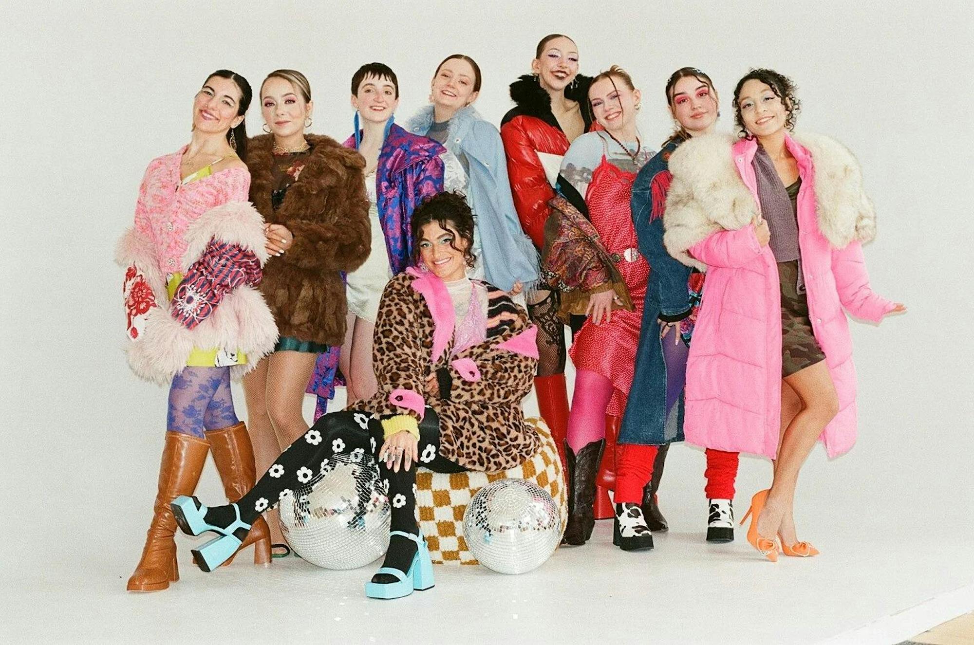 9 young adults pose for a photoshoot wearing elaborate outfits featuring bright colors and mixed patterns. The group stands behind Chloe Gray, who sits on a cushion in front of them.