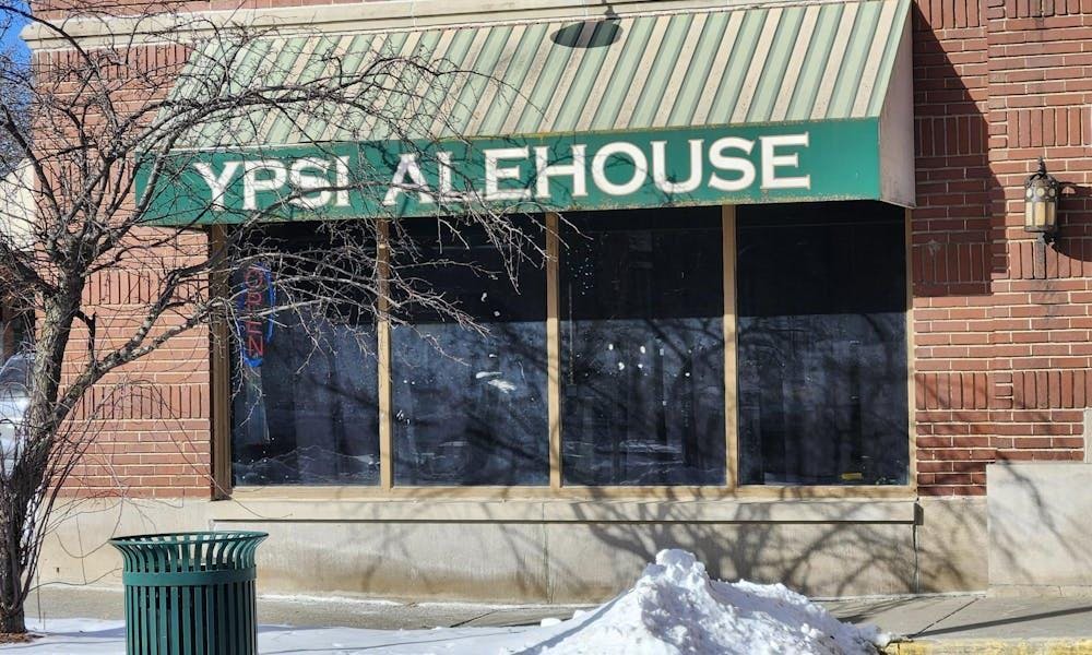 The exterior of a red brick building with a green awning over a large window. The awning says "Ypsi Alehouse" in capital white letters. In front of the building are a pile of white snow and a tree that has lost all its leaves. 